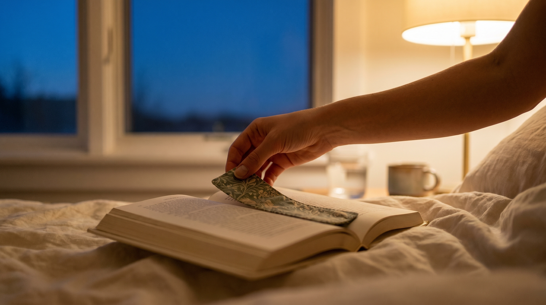 A hand placing a fabric bookmark in an open book on cream linen with warm bedside lamp